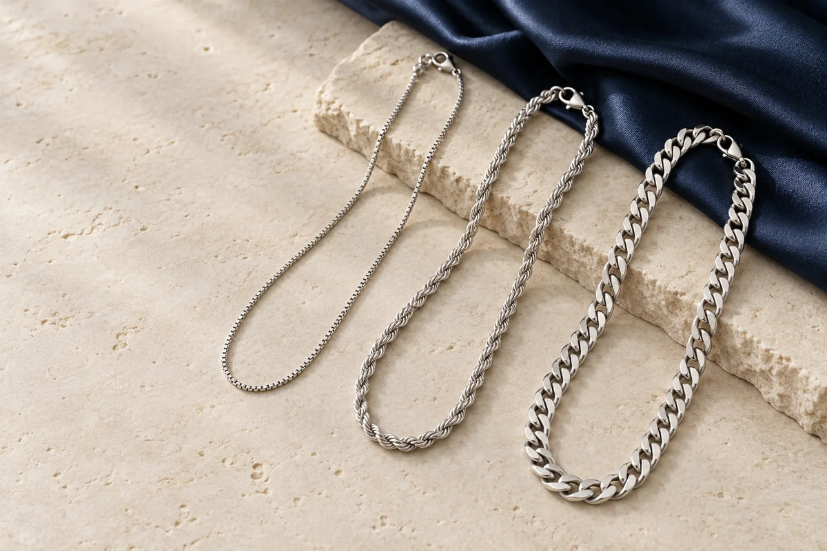 Illustrative styling arrangement of silver box, rope and flat curb chains on ivory stone and navy fabric